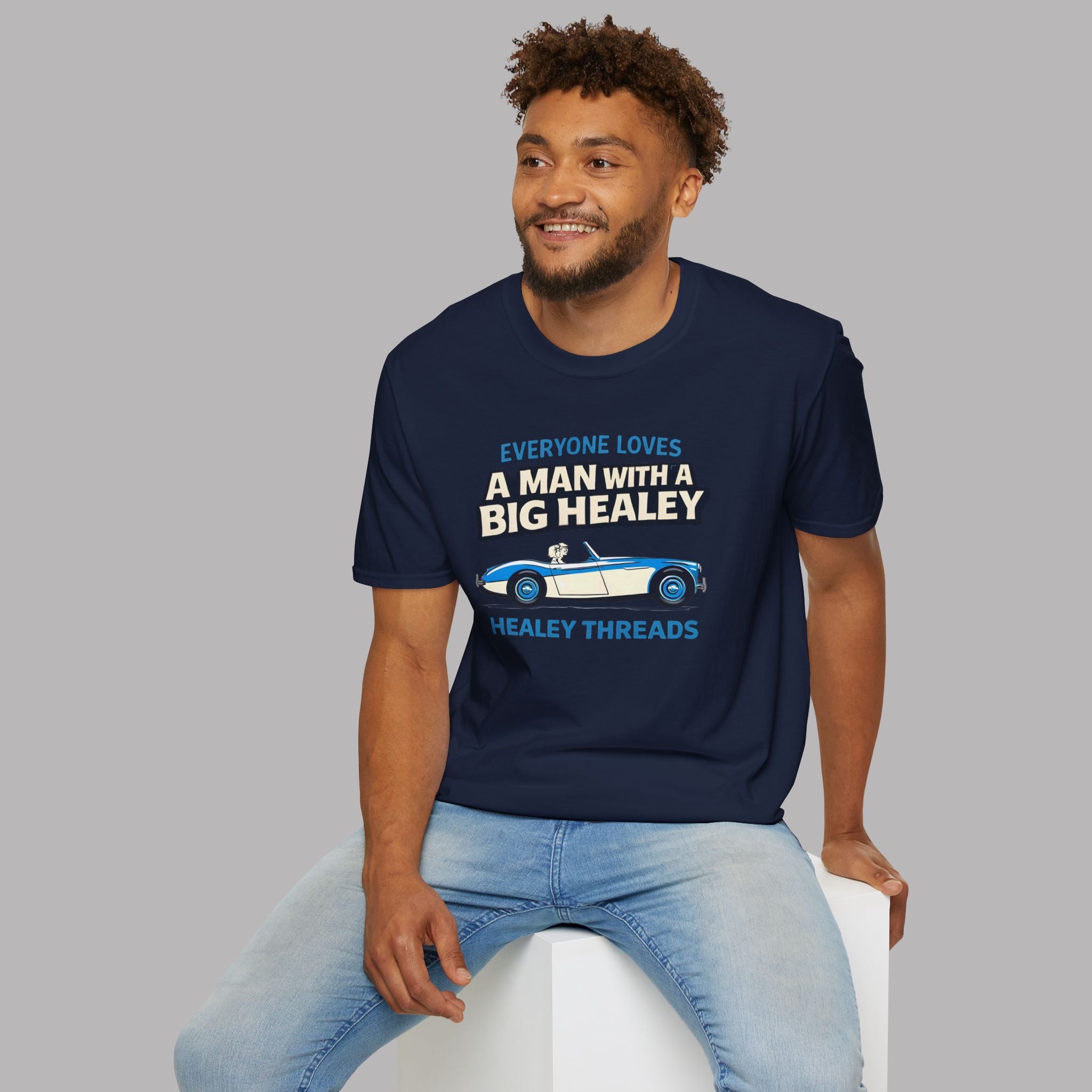 Man wearing a navy blue t-shirt with text and Healey car graphic, sitting on a white stool against a gray background.