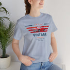 Woman wearing a light blue t-shirt with a vintage car design, standing indoors next to a plant.