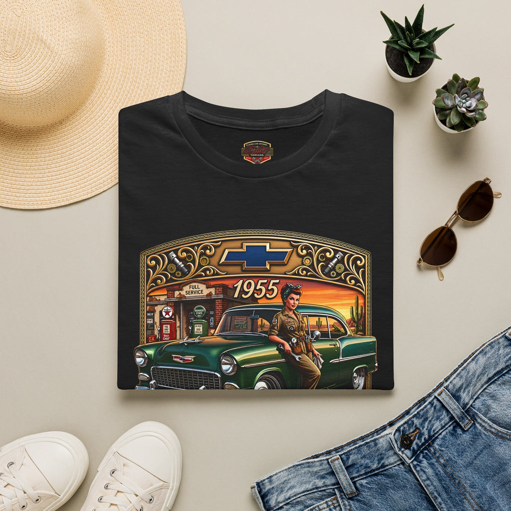 A folded black graphic T-shirt is displayed in a styled flat lay on a light beige surface, surrounded by a straw hat, white sneakers, blue jeans, small potted succulents, and sunglasses. The shirt features a large vintage-style print of a green classic car, a woman in military attire, a gas station, and the Chevrolet bowtie logo with '1955' text.