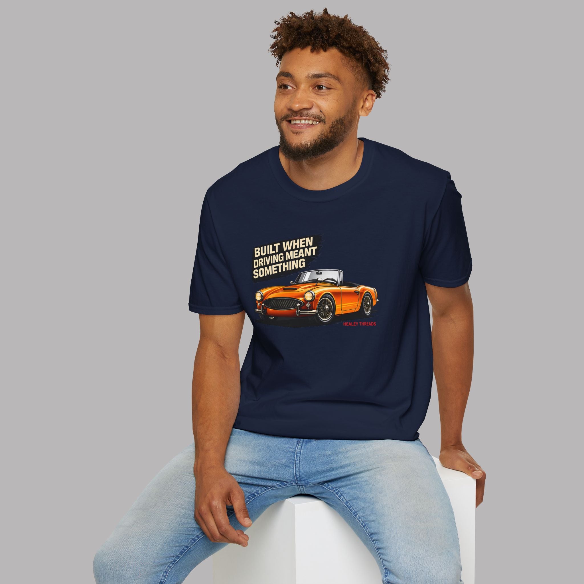 Man wearing a navy blue t-shirt with a graphic and text, sitting on a white stool against a gray background.