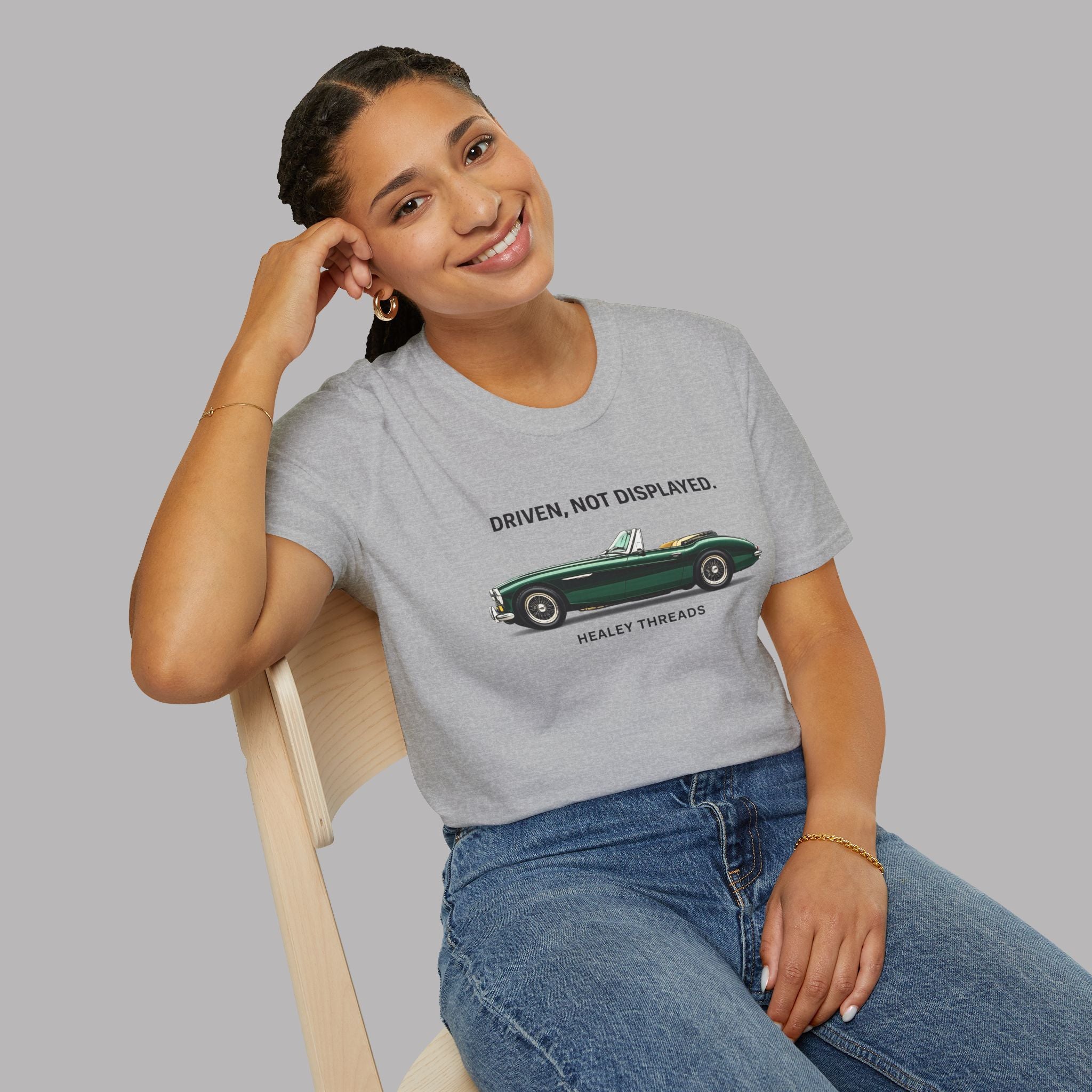Woman wearing a gray t-shirt with a vintage car graphic and text, sitting on a chair against a plain background.