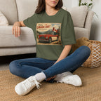 A woman sitting cross-legged on a woven rug in front of a light beige sofa, wearing an military green graphic t-shirt with a vintage-style 'Welcome to Canada Day!' print featuring a red car and Canadian flag.