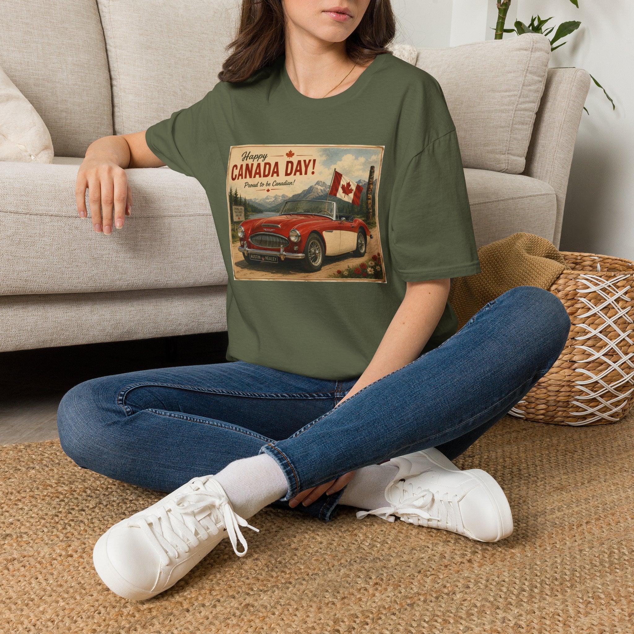 A woman sitting cross-legged on a woven rug in front of a light beige sofa, wearing an military green graphic t-shirt with a vintage-style 'Welcome to Canada Day!' print featuring a red car and Canadian flag.