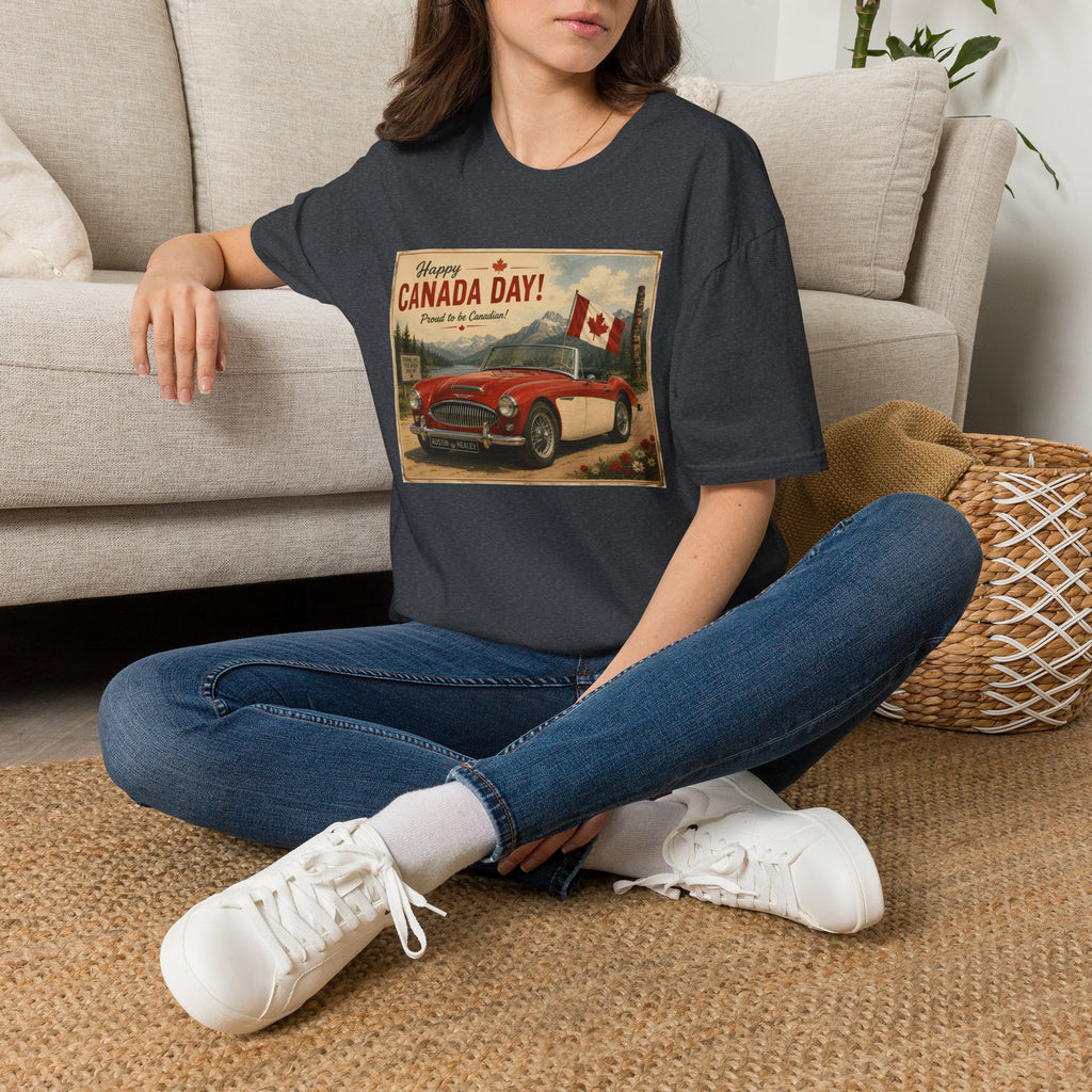 A woman sitting cross-legged on a woven rug in front of a light beige sofa, wearing a dark charcoal gray graphic t-shirt with a vintage-style 'Welcome to Canada Day!' print featuring a red car and Canadian flag.