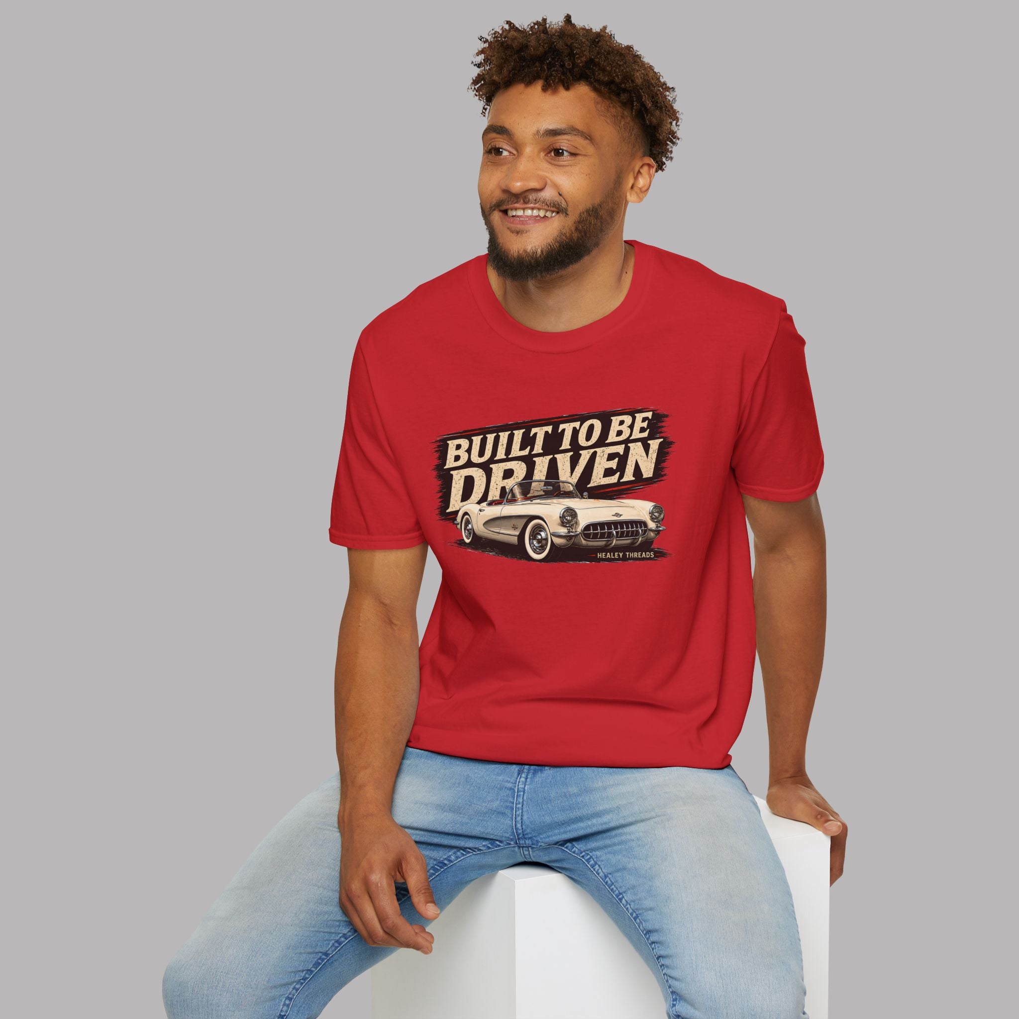 Man wearing a red t-shirt with a built to be driven graphic and text, sitting on a white stool against a gray background