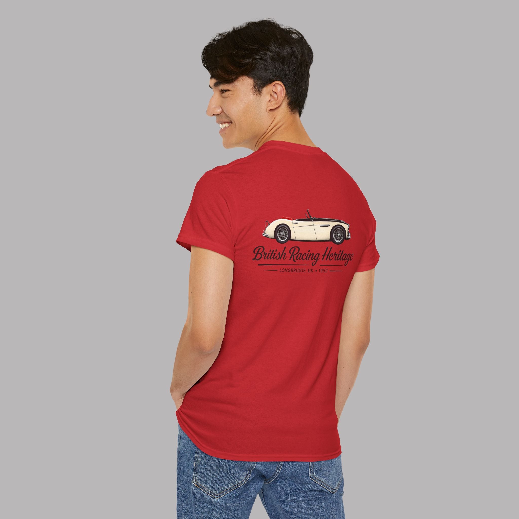 Back view of a man wearing a red t-shirt with a Healey car design and black text on the back, standing against a gray background