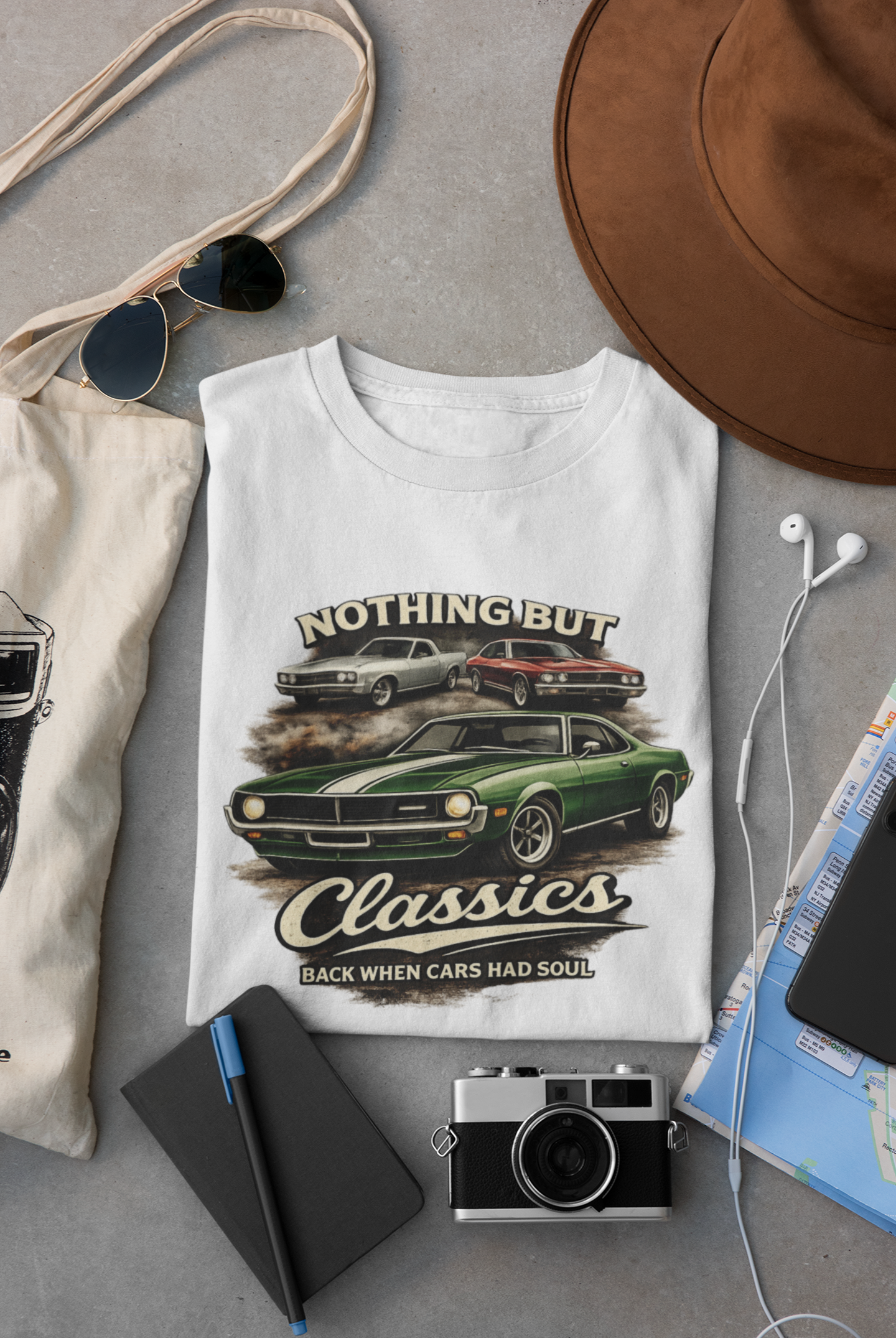 White t-shirt with classic car graphic and text, surrounded by sunglasses, hat, camera, and map on a gray surface.