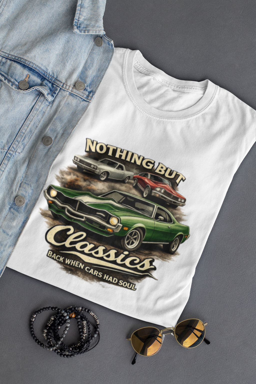 White t-shirt with car graphic and text, denim jacket, sunglasses, and bracelets on a gray background
