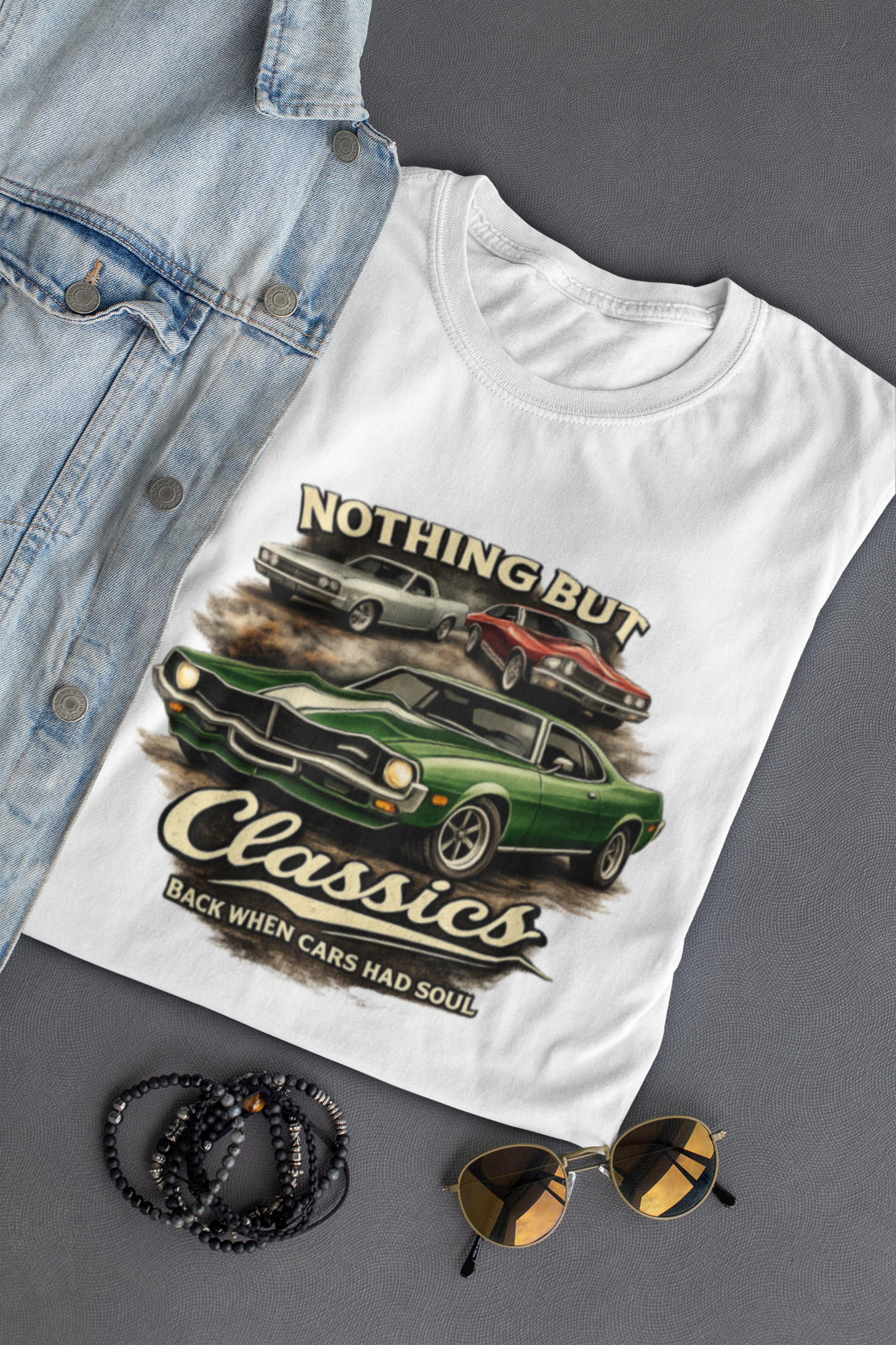 White t-shirt with car graphic and text, denim jacket, sunglasses, and bracelets on a gray background