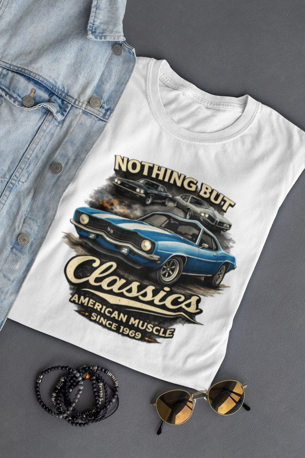 White t-shirt with car graphic and text on a gray surface with denim jacket, sunglasses, and bracelets.