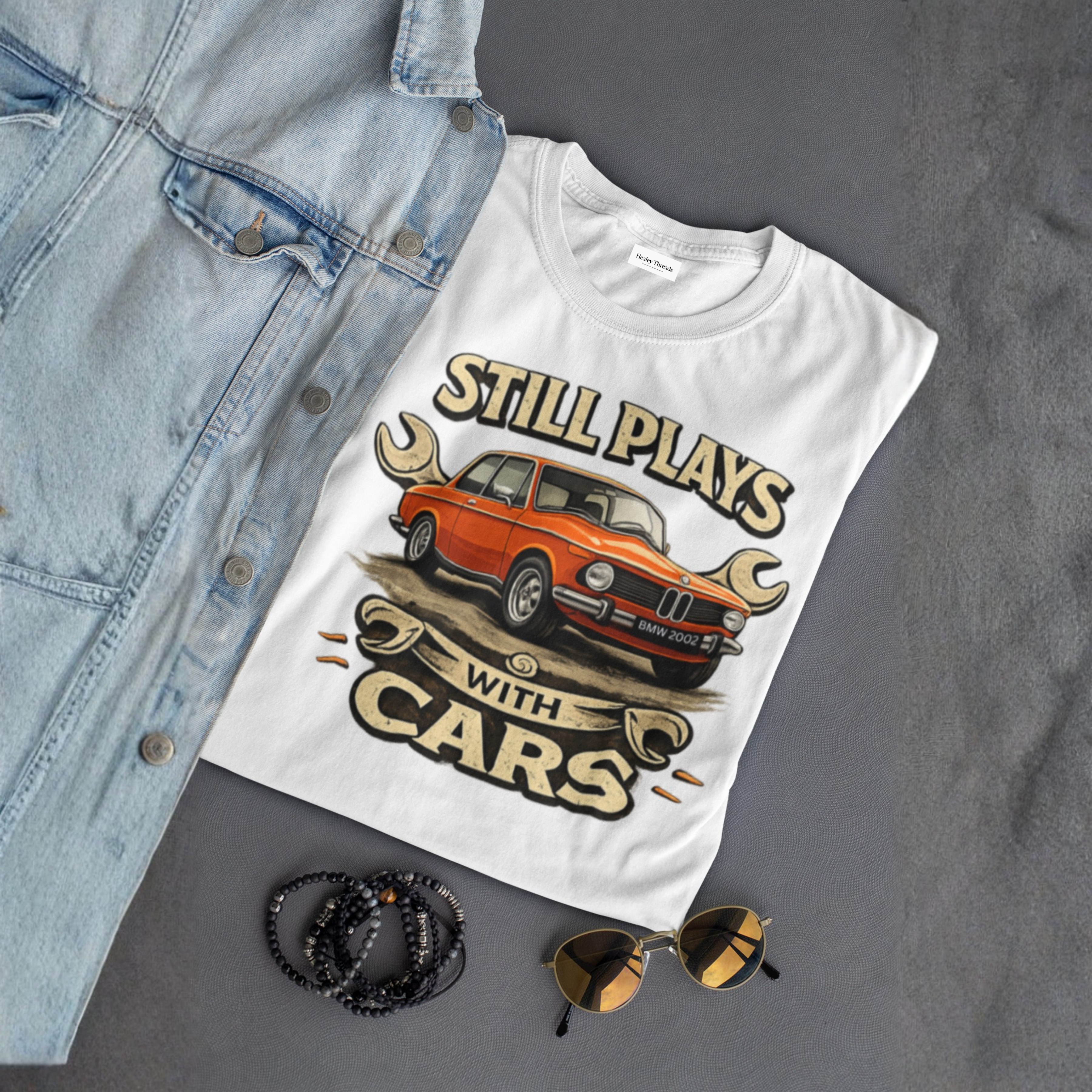 White t-shirt with car graphic and text 'Still Plays with Cars' on a gray surface with denim jacket, sunglasses, and bracelets.