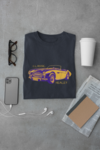 Blue t-shirt with a purple and yellow classic car design on a gray surface with a phone, glasses, and newspaper.