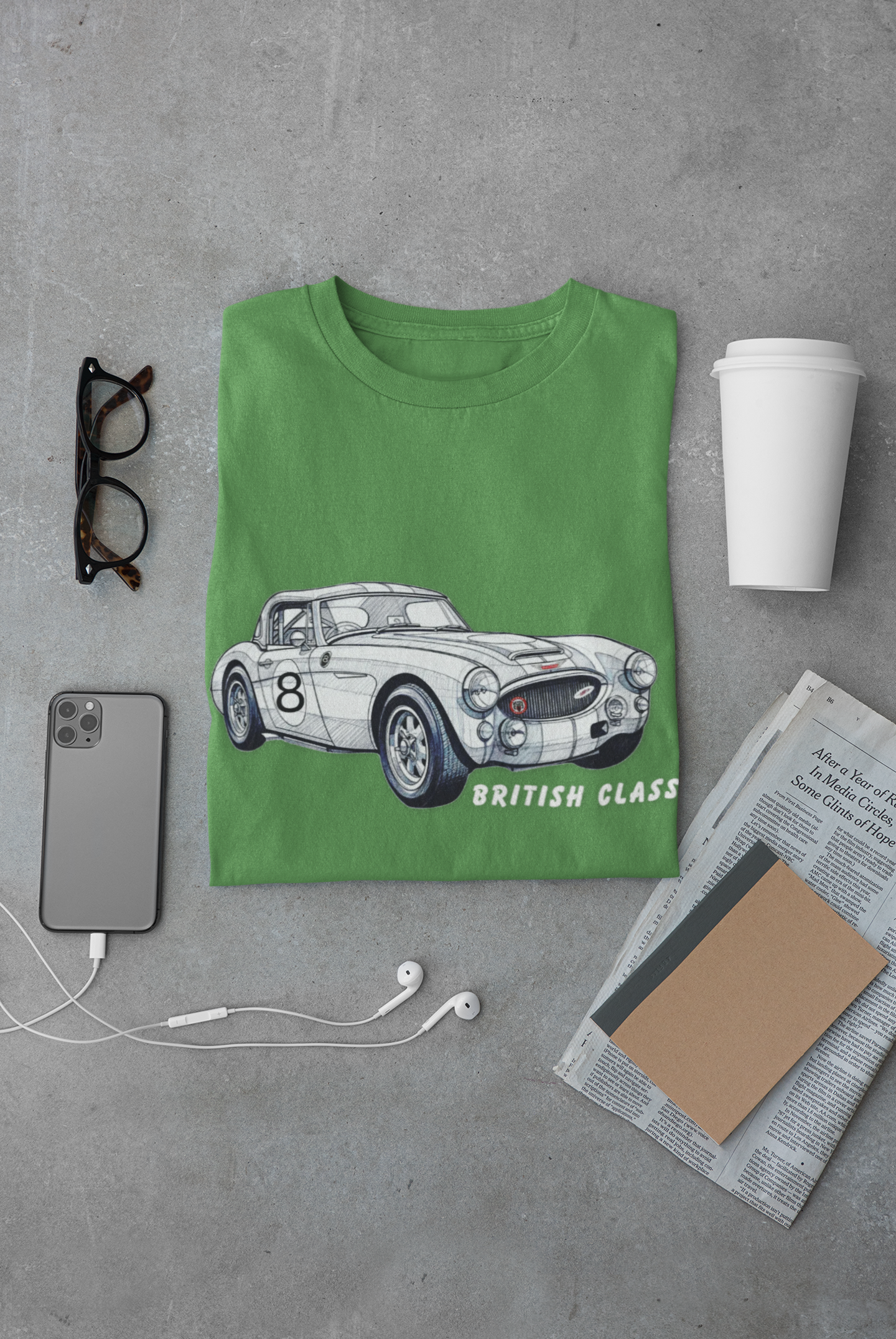Green t-shirt with a car graphic and 'British Classic' text on a gray surface with a phone, glasses, and newspaper.