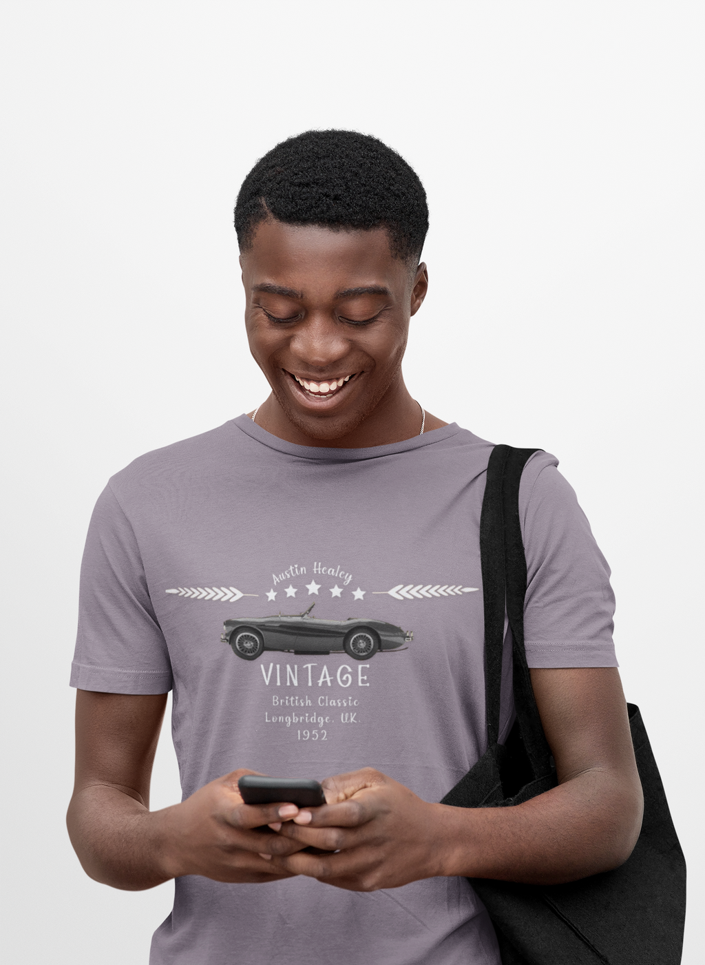 Man wearing a gray t-shirt with a vintage car design, holding a phone and a black bag on a light gray background