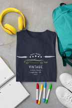 Navy blue t-shirt with vintage car design on a gray surface with headphones, backpack, and markers.