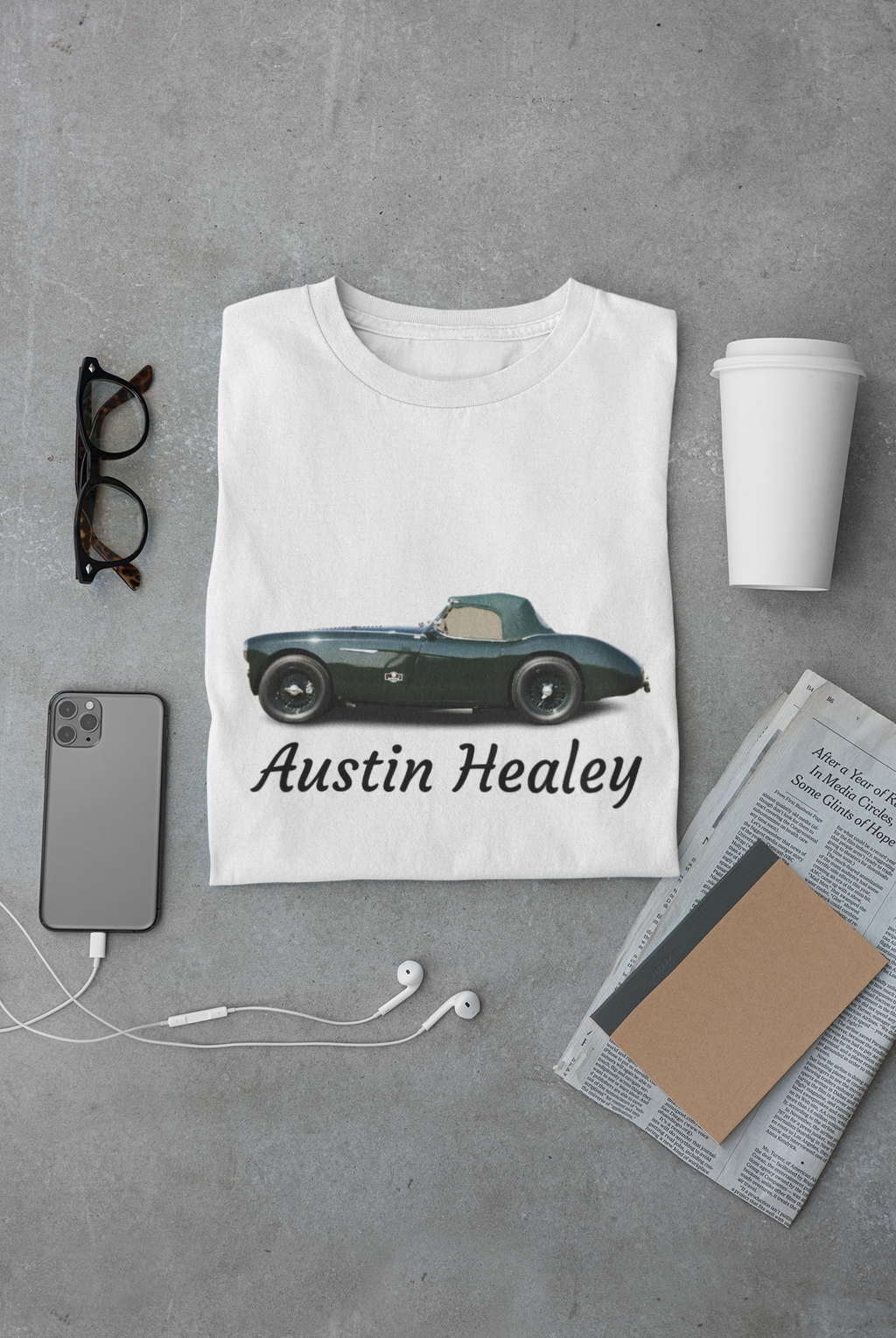 White t-shirt with Austin Healey car design on a gray surface with phone, glasses, and cup.