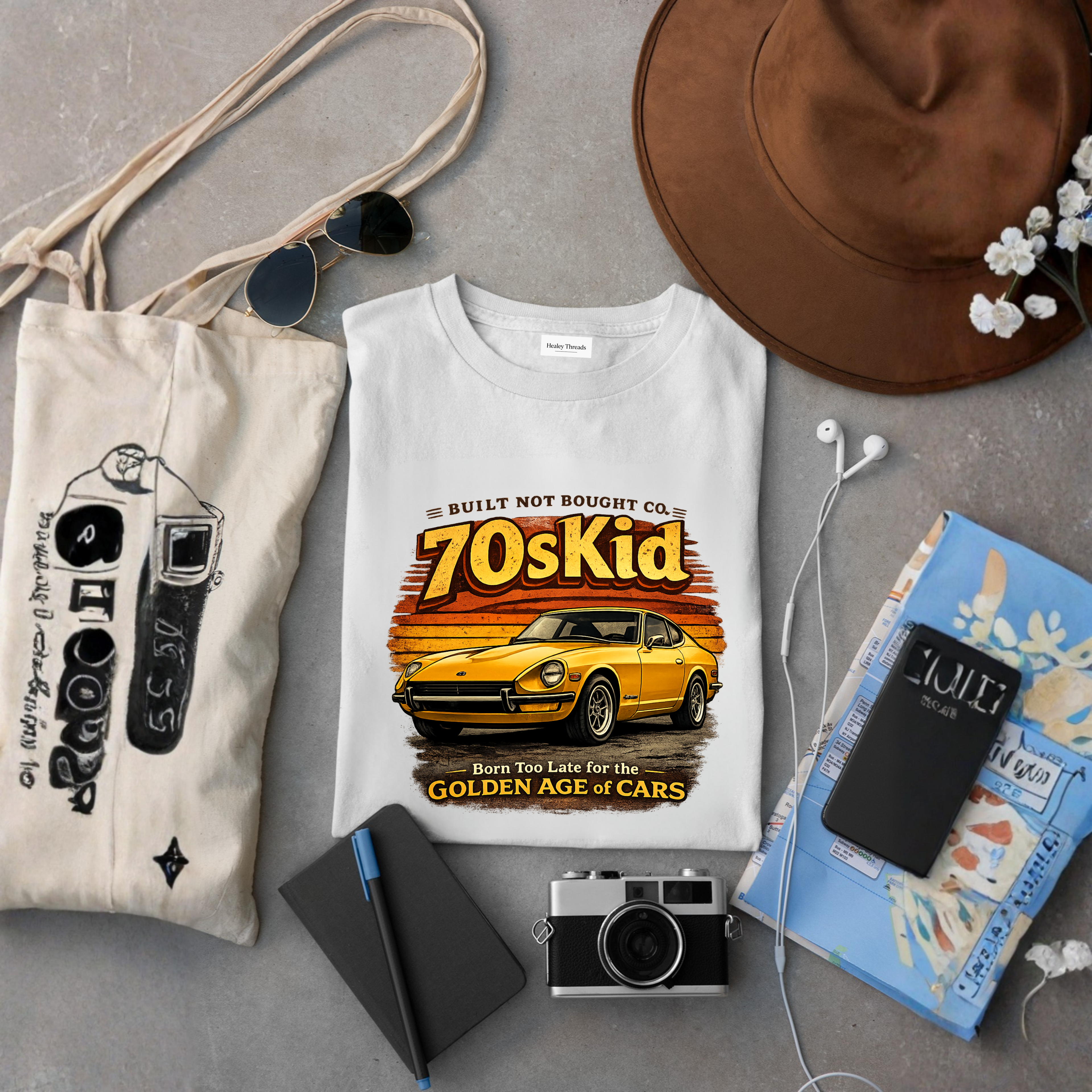 Folded white t-shirt with a yellow vintage car and “70s Kid – Born Too Late for the Golden Age of Cars” design, surrounded by a hat, tote bag, sunglasses, notebook, camera, phone, and earphones.