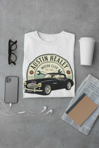 White t-shirt with Austin Healey logo on a gray surface with phone, cup, and glasses.