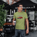 Man wearing a green t-shirt with a graphic and text, standing outdoors under a Deluca restaurant umbrella.