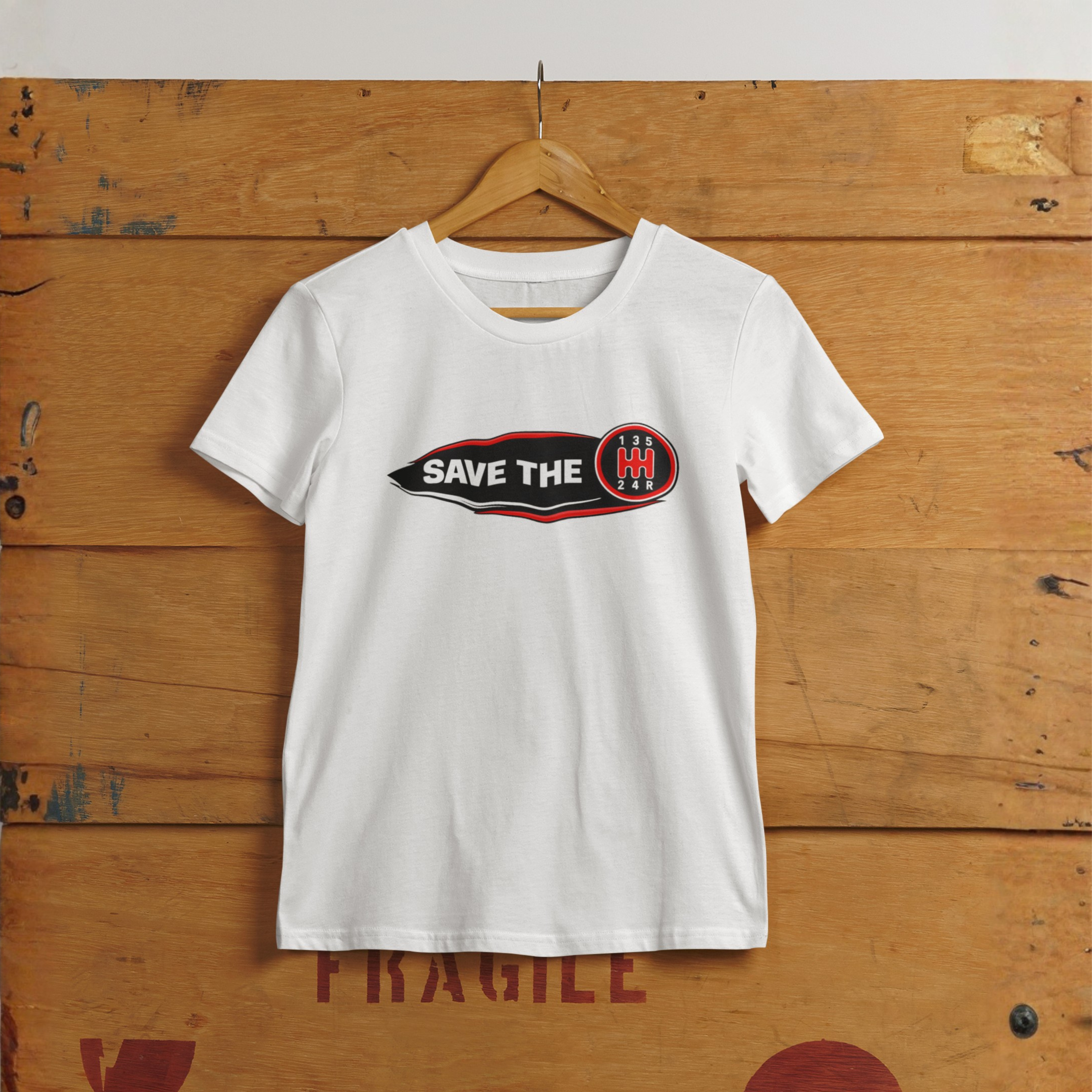 A white t-shirt with 'Save the stick shift' graphic design hanging on a wood wall.