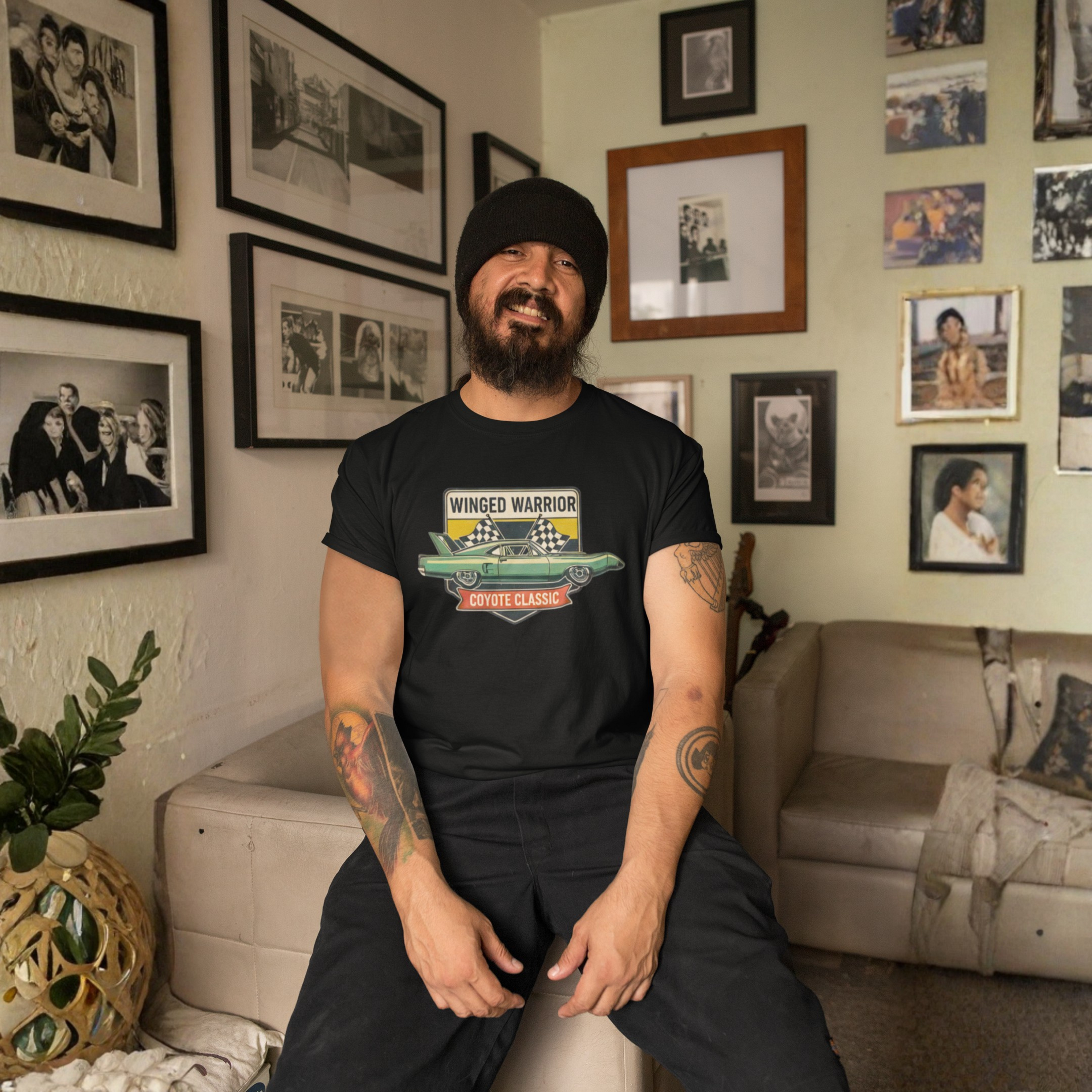 Man wearing a black t-shirt with a graphic design, sitting in a living room with framed photos on the wall.