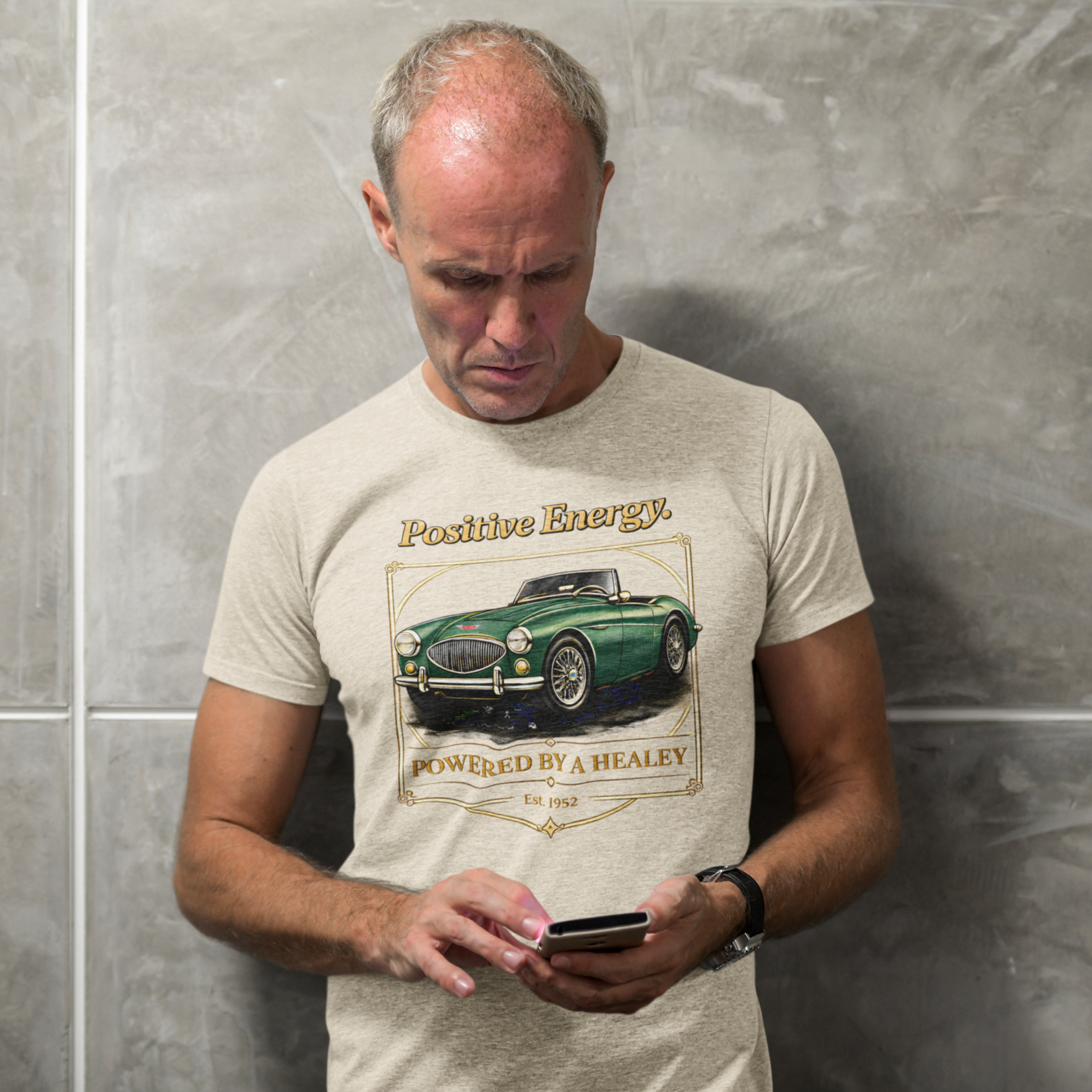 Man wearing a sand colored t-shirt with a vintage car graphic and text, standing against a gray wall.