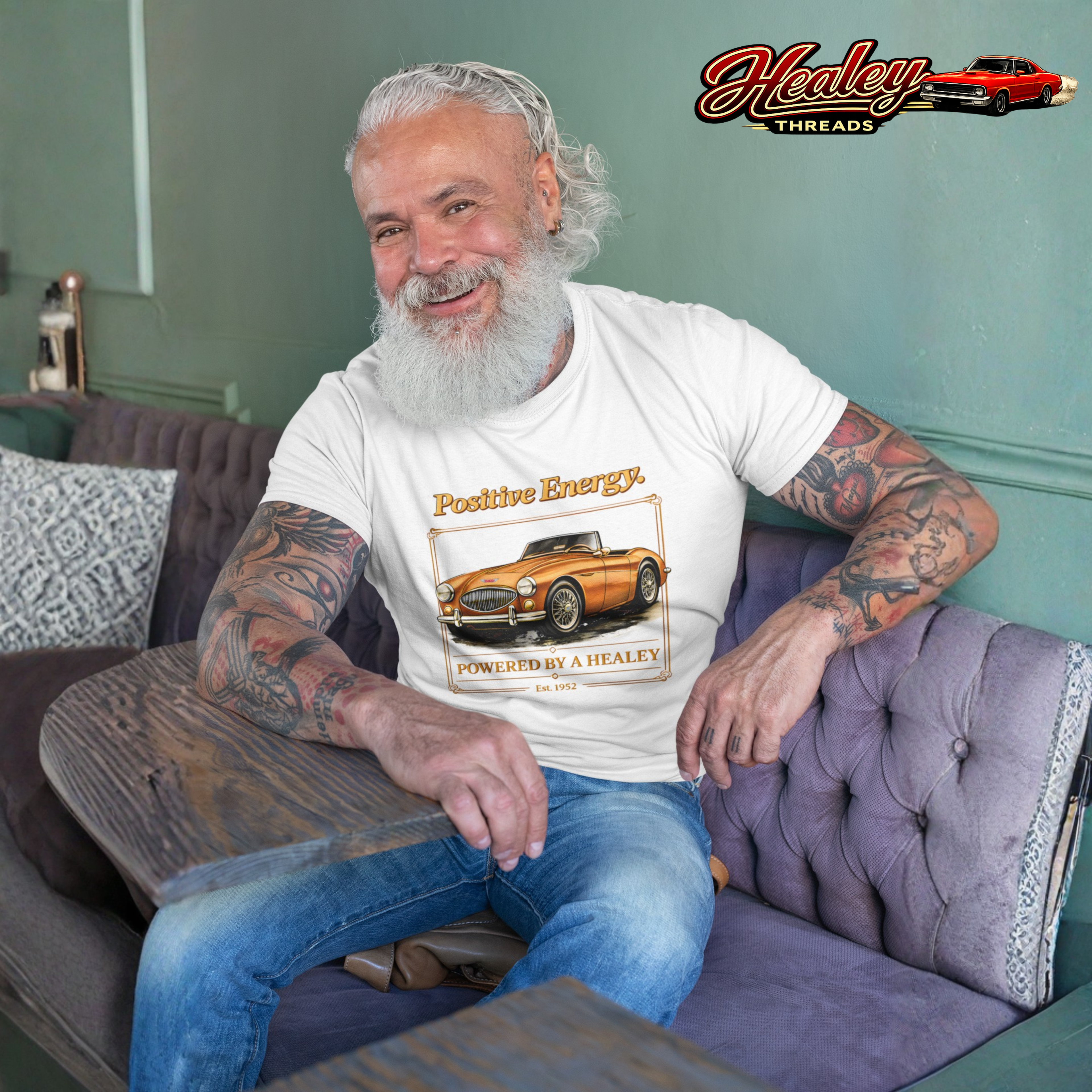 Man sitting on a couch wearing a white t-shirt with a classic car graphic and 'Healey Threads' logo.