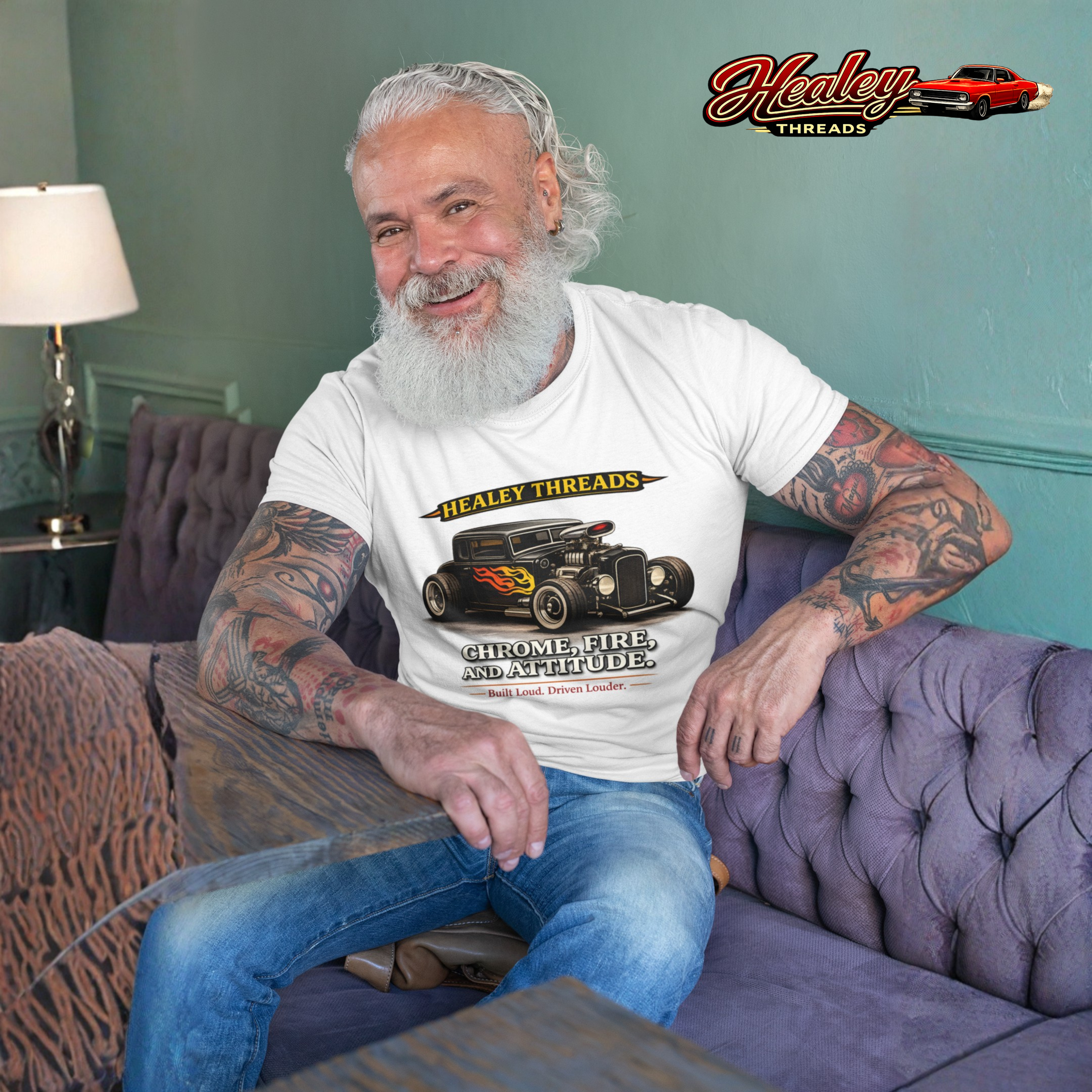 Man sitting on a couch wearing a white t-shirt with a graphic hot rod car design, in a room with a lamp and decorative elements.