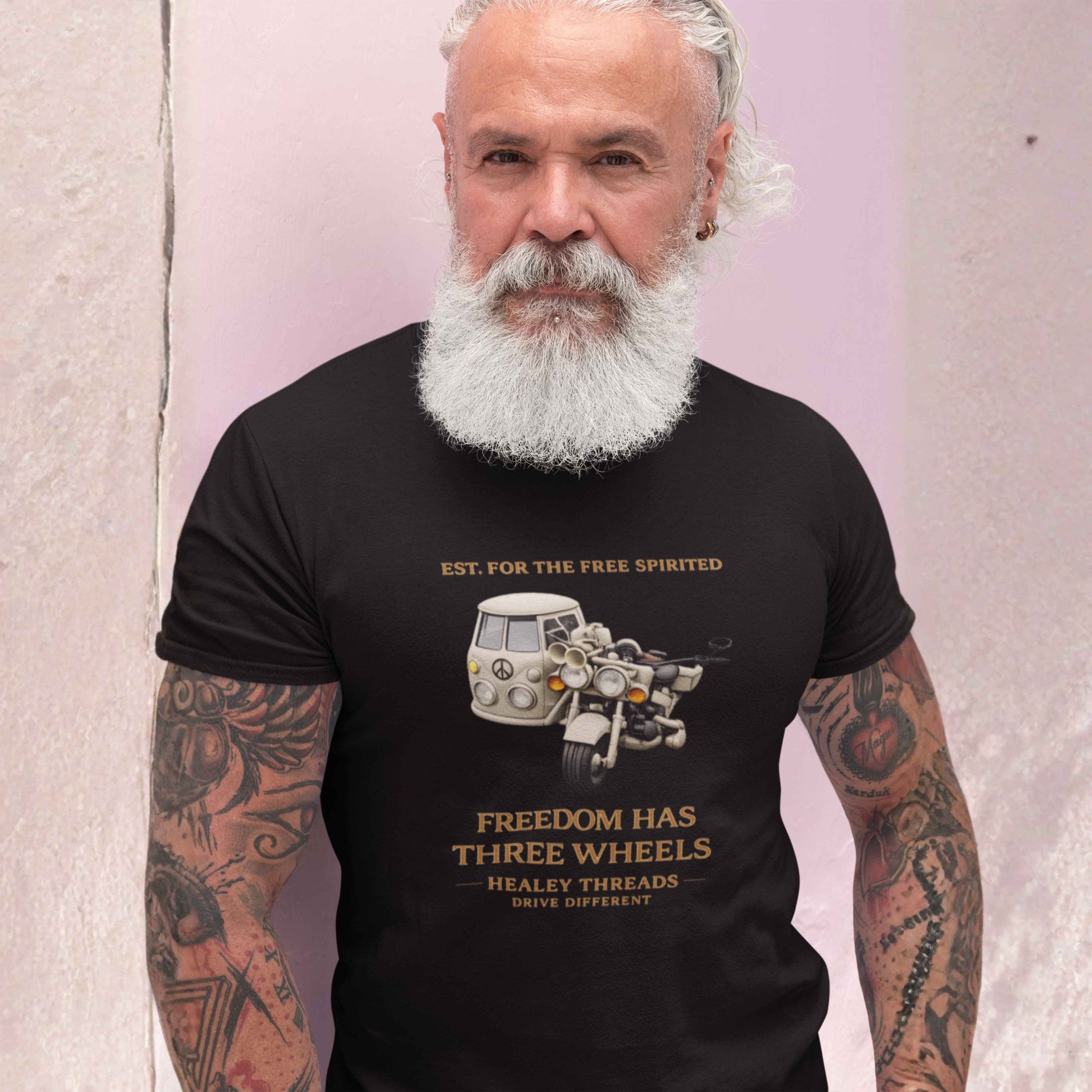 Man wearing a black t-shirt Cream motorcycle-sidecar van mashup with peace symbol and gold "Freedom Has Three Wheels" text, standing against a light pink wall.