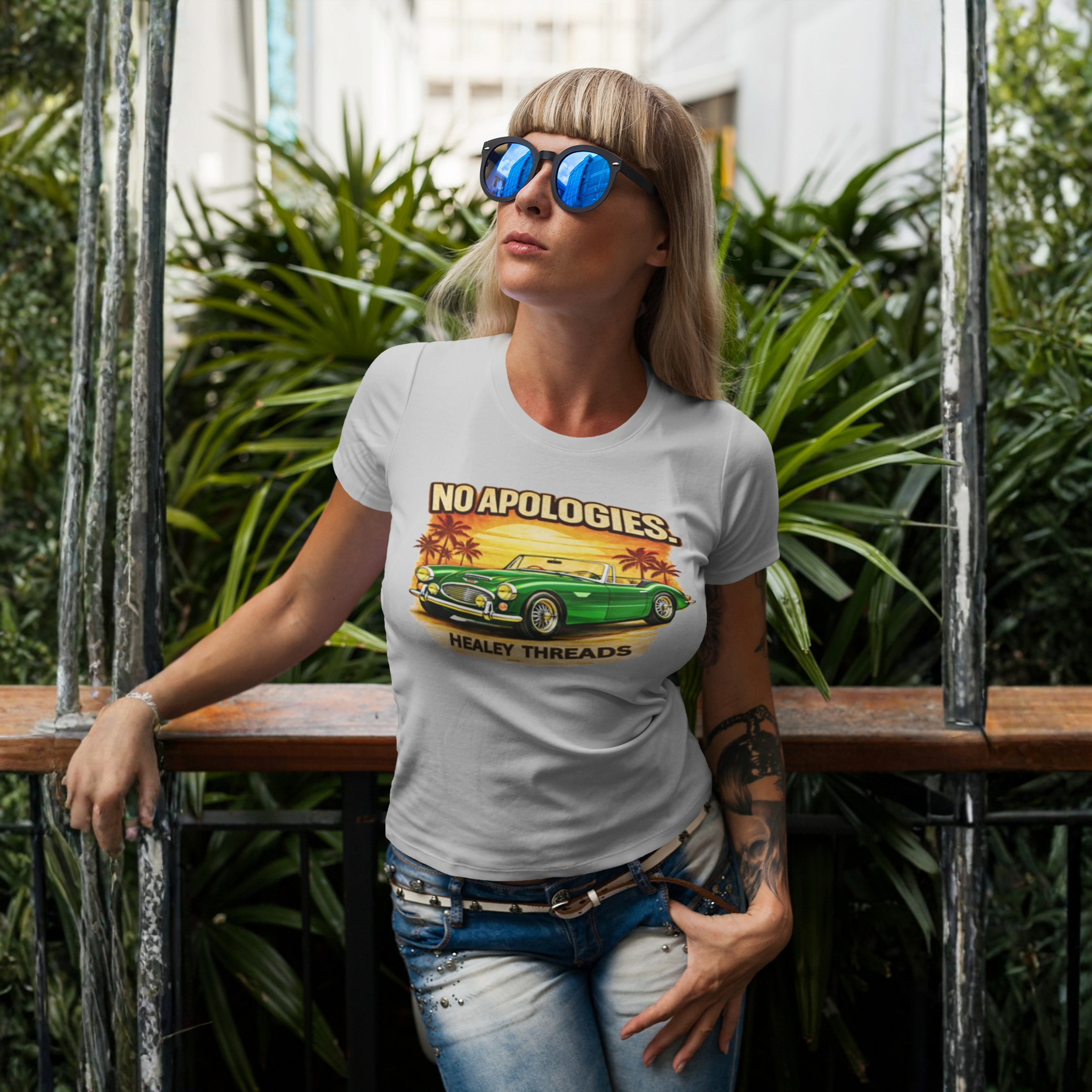 Woman wearing a white t-shirt with a vintage Healey car graphic and text, standing outdoors with plants in the background.