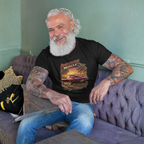 Man with tattoos and a beard sitting on a purple couch wearing an 'American Muscle' t-shirt.