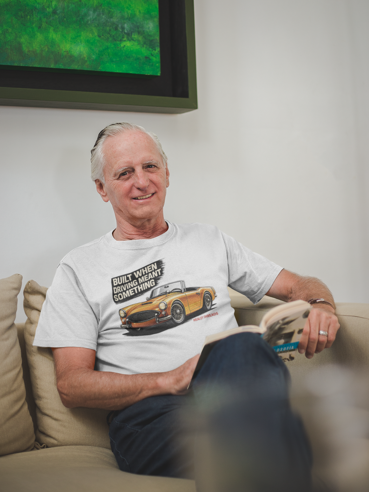 Man sitting on a couch wearing a white t-shirt with a British classic car graphic design, smiling.