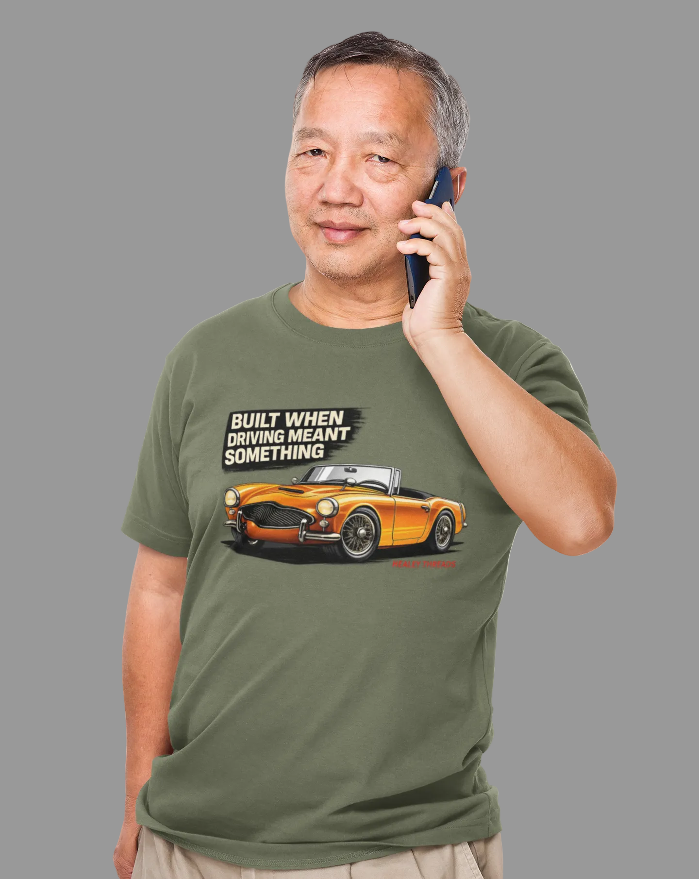 Man wearing a military green t-shirt with a British car graphic and text, talking on a phone.