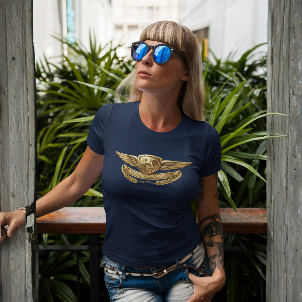Woman wearing a navy blue t-shirt with a gold Healey emblem, standing outdoors with greenery in the background.