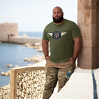 Man wearing a military green t-shirt with a Austin Healey logo, standing by a coastal wall.