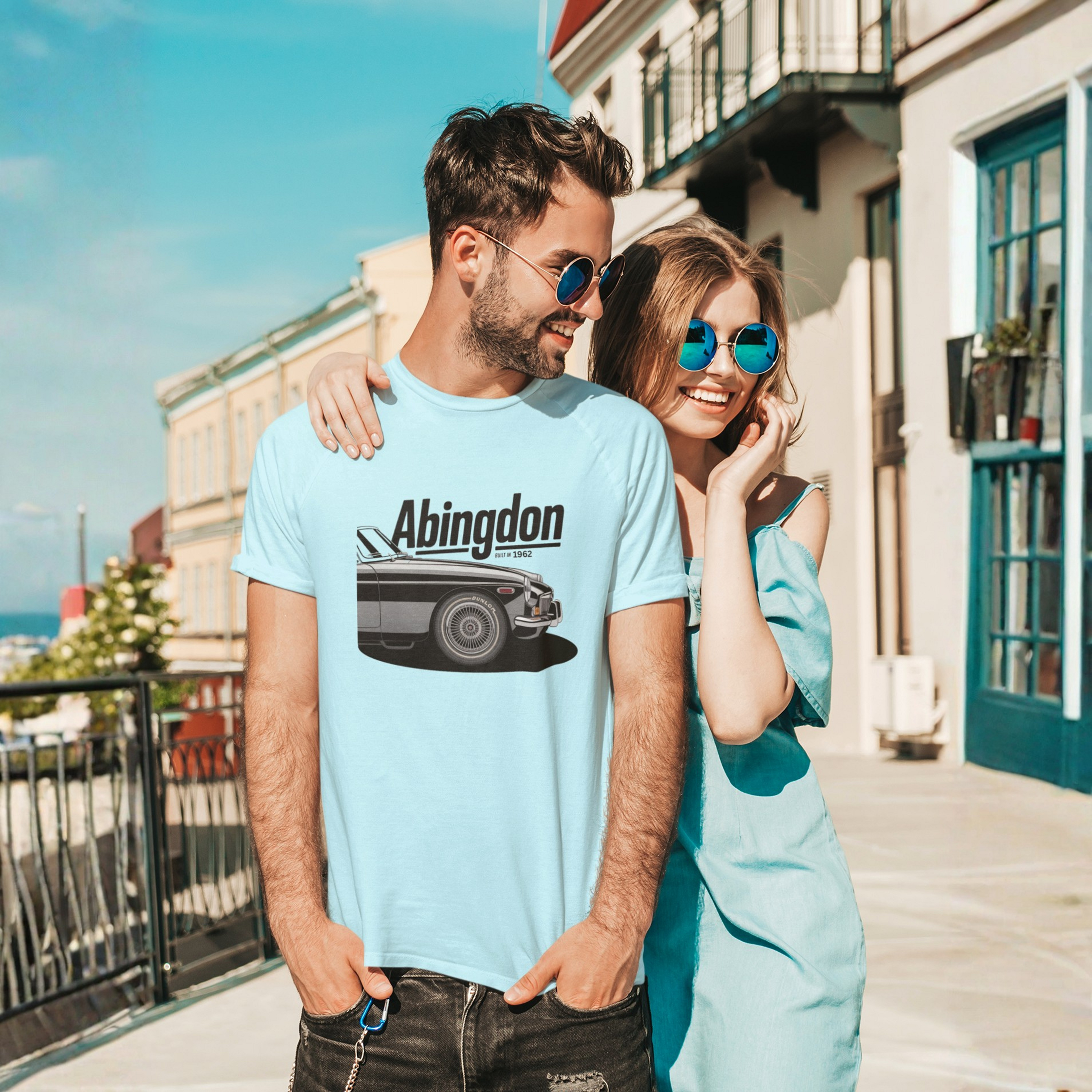Man and woman standing together on a sunny day, both wearing light blue shirts with a classic car graphic and 'Abingdon' text.