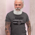 Man wearing a dark heather t-shirt with 'Abingdon' and classic car graphic against a light pink background