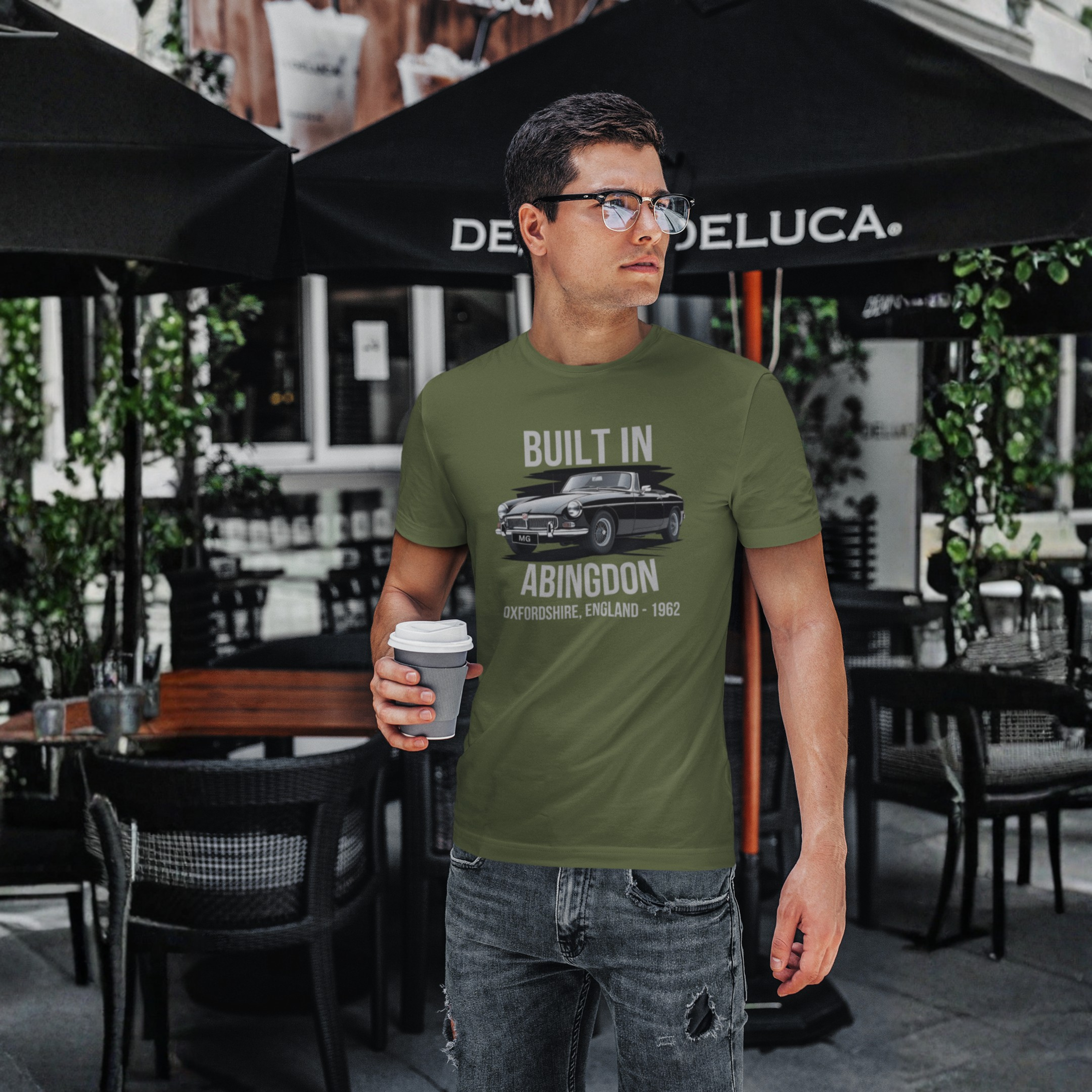 Man wearing a military green t-shirt with text and a classic car graphic, holding a coffee cup outdoors.
