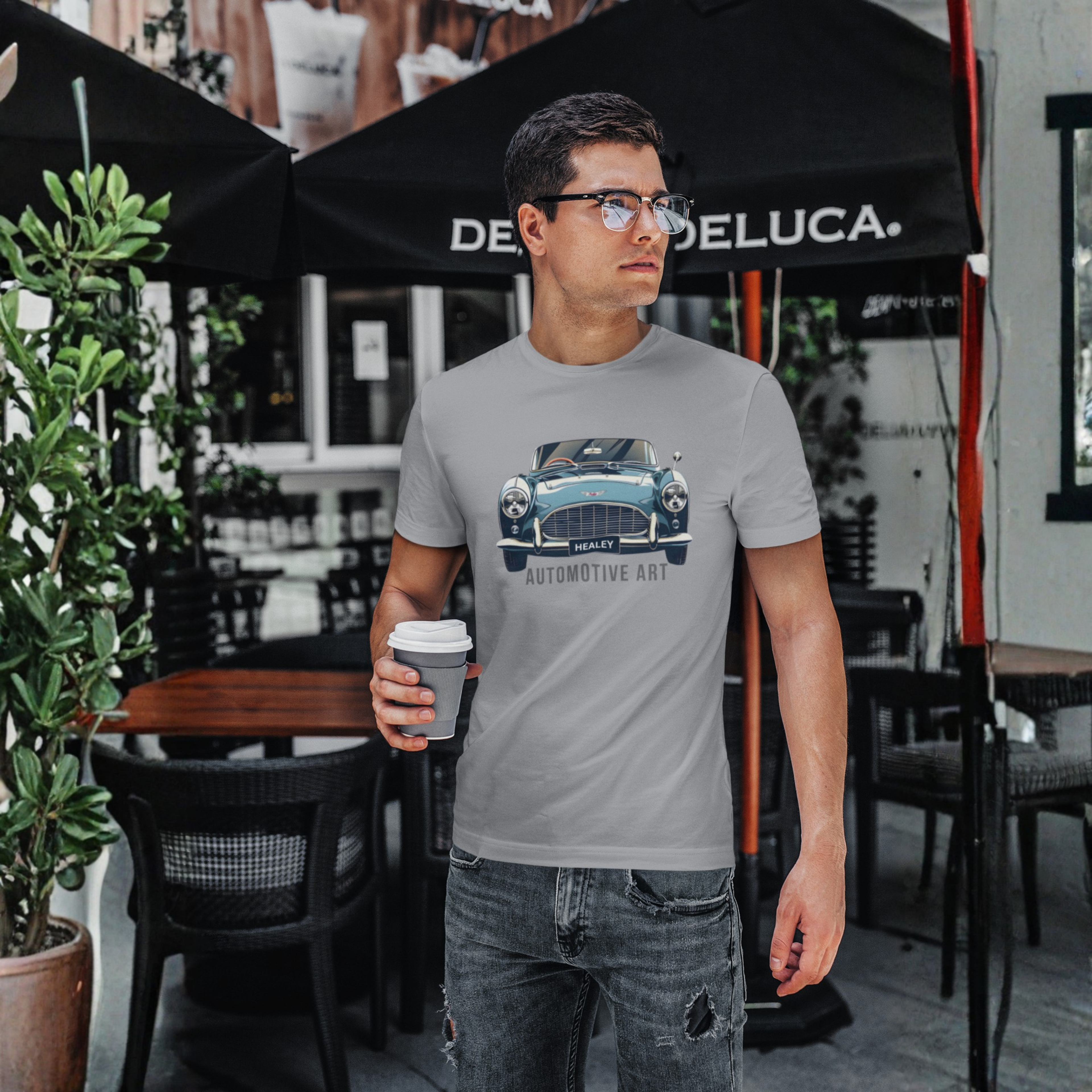 Man wearing a sport gray t-shirt with a Healey car design, holding a coffee cup, standing in front of a Deluca store.