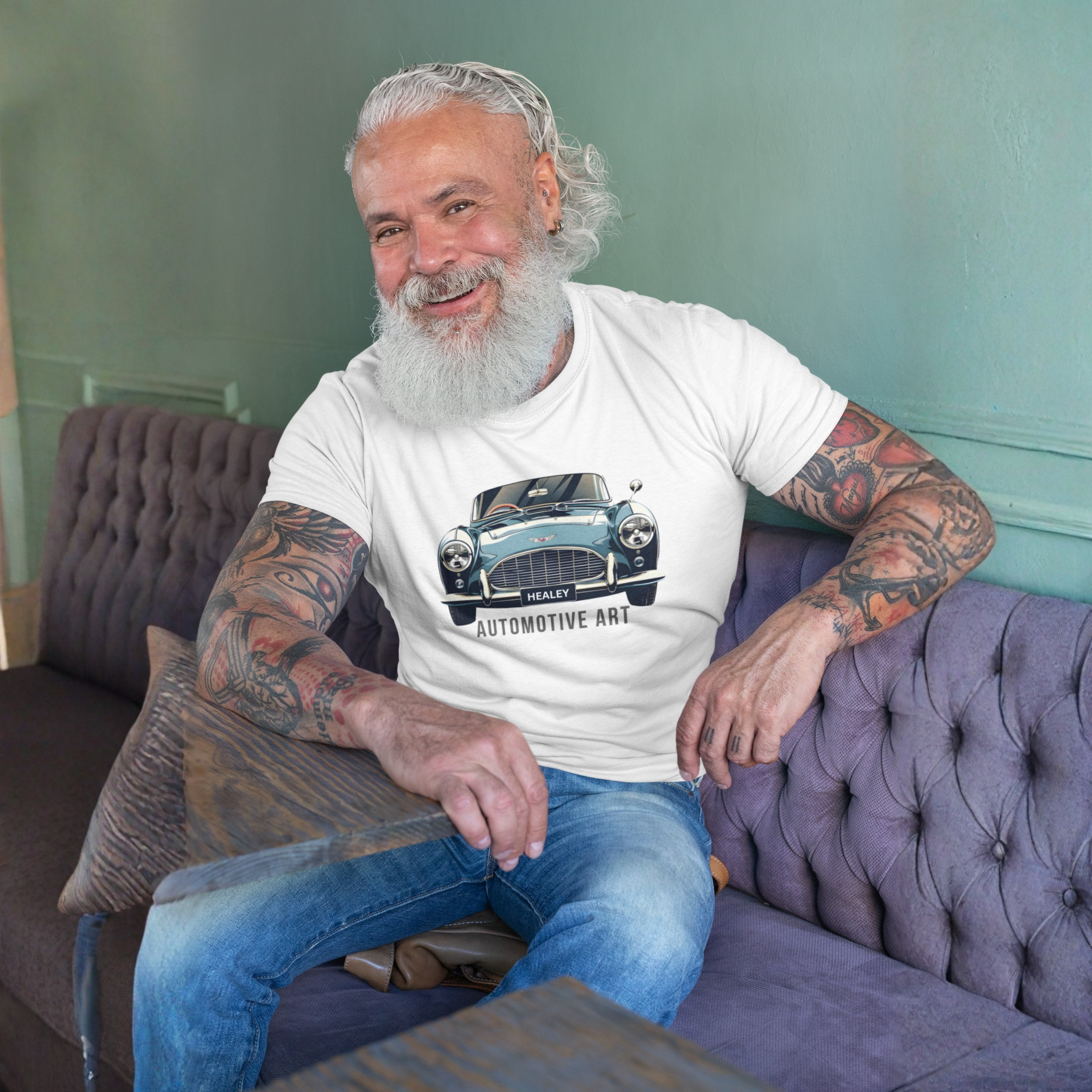 Man with tattoos sitting on a purple couch wearing a white t-shirt with a Healey car design.