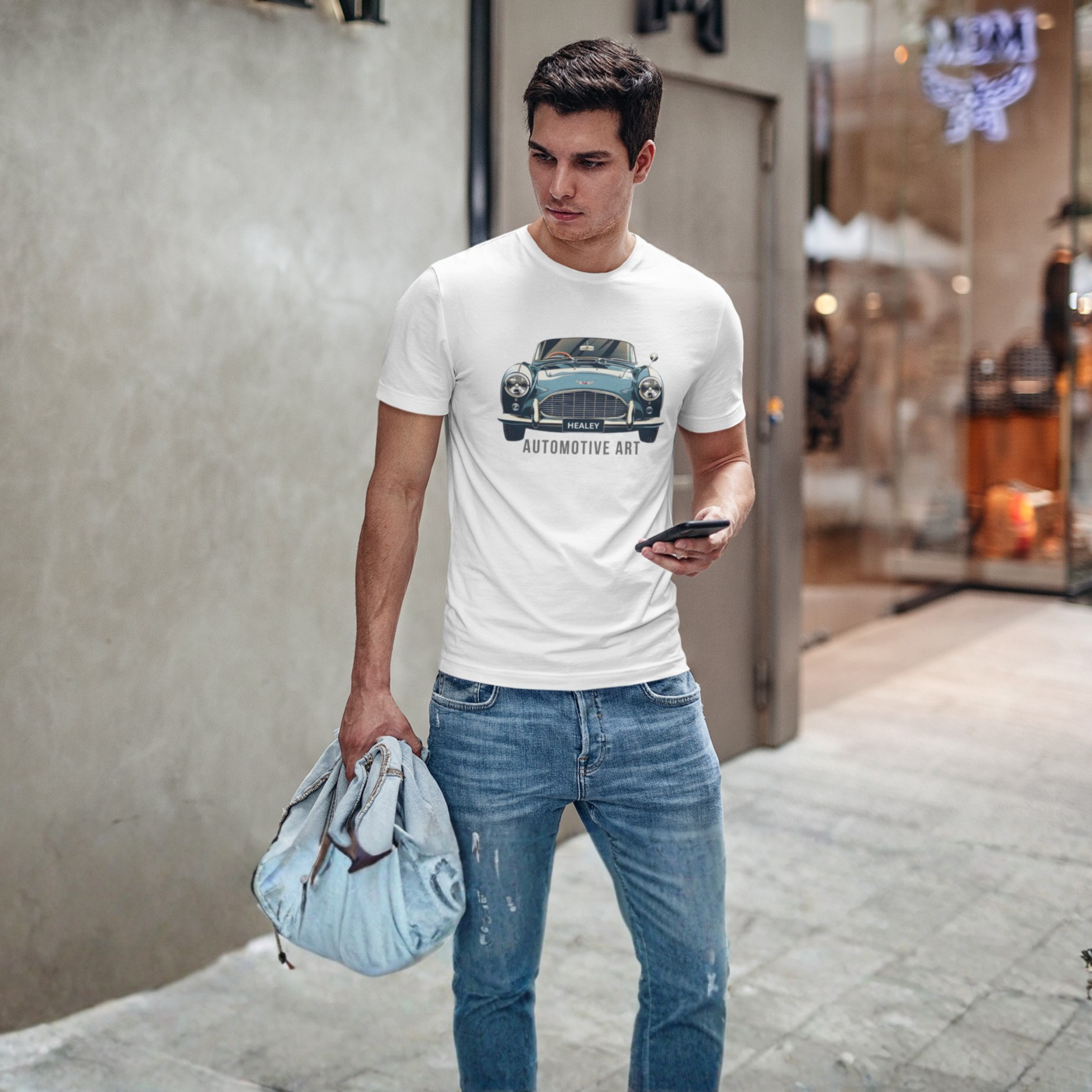 Man wearing a white t-shirt with a Healey car graphic and blue jeans, holding a bag and phone, standing on a street.e