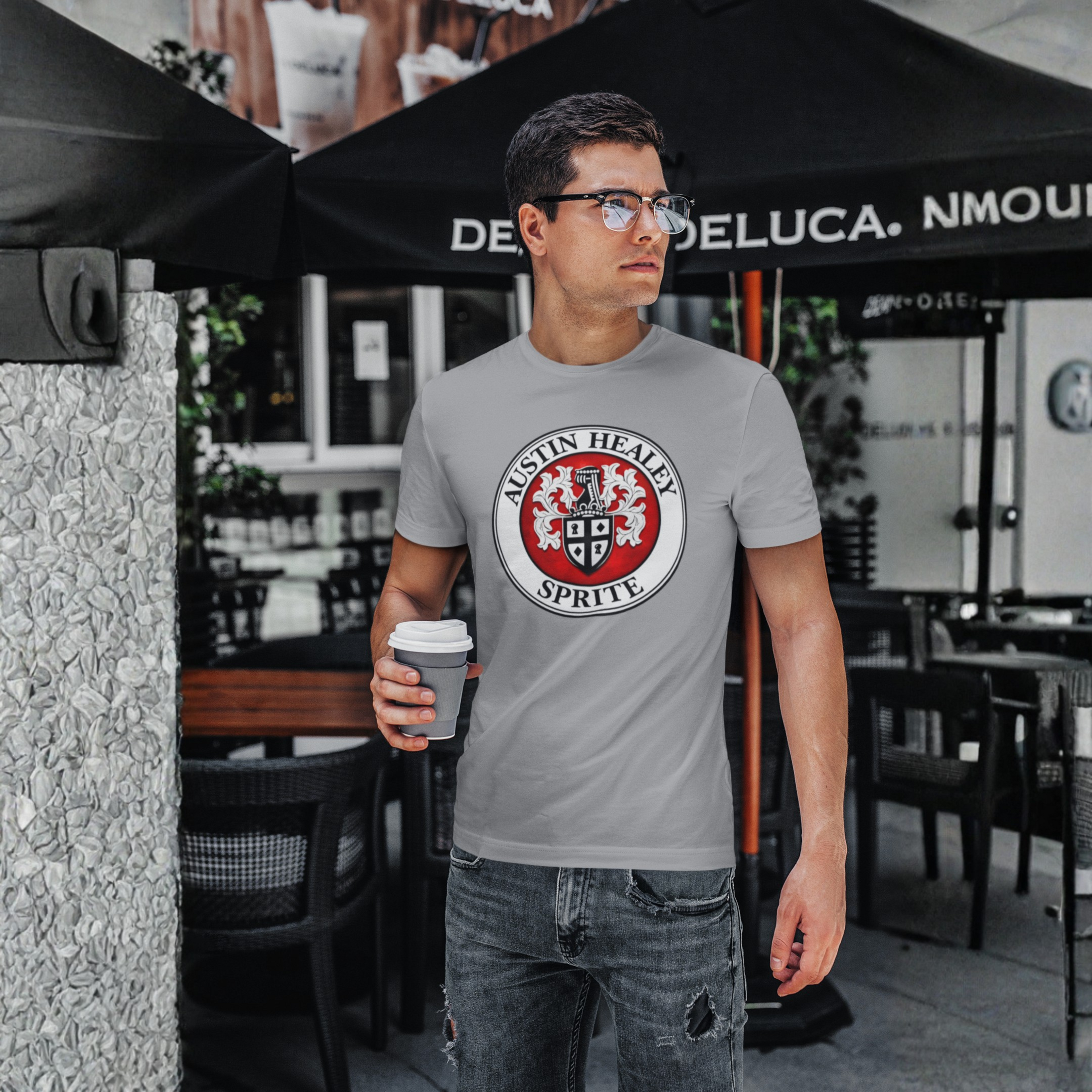 Man wearing a gray t-shirt with a Austin Healey Sprite logo, holding a coffee cup, standing outdoors.