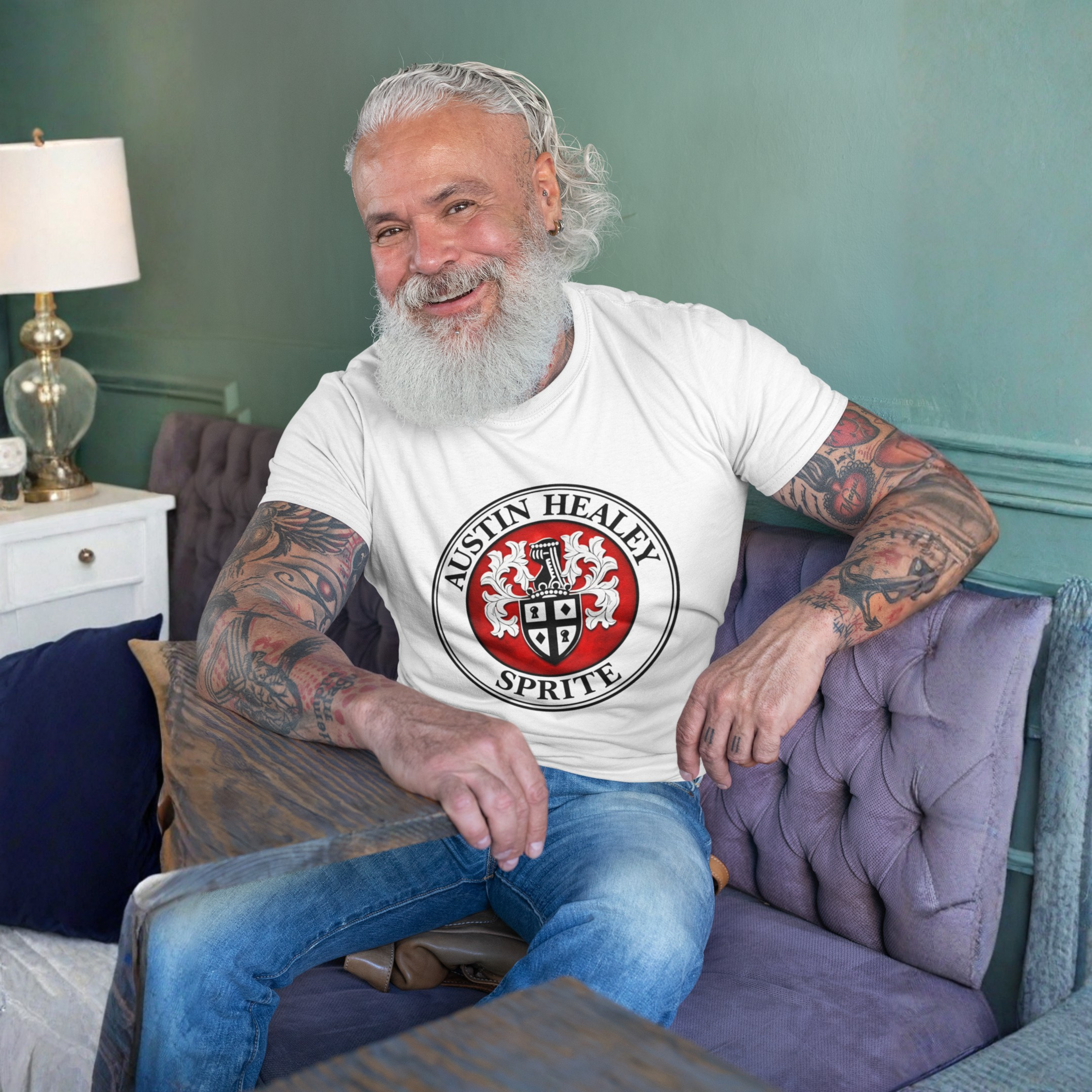 Man sitting on a couch wearing a white t-shirt with a red and black Austin Healey Sprite logo, sitting in a room with a lamp and nightstand.