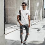 Man wearing a white t-shirt with a Healey design on left chest and ripped jeans walking outdoors.