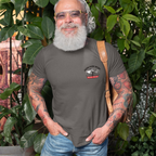 Man wearing a gray t-shirt with a logo on left chest, standing outdoors among greenery.