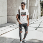 Man wearing a white t-shirt with a graphic Healey car design, ripped jeans, and carrying a backpack on a city street.