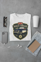 White t-shirt with 'Healey Threads' design on a gray surface with phone, cup, and glasses.