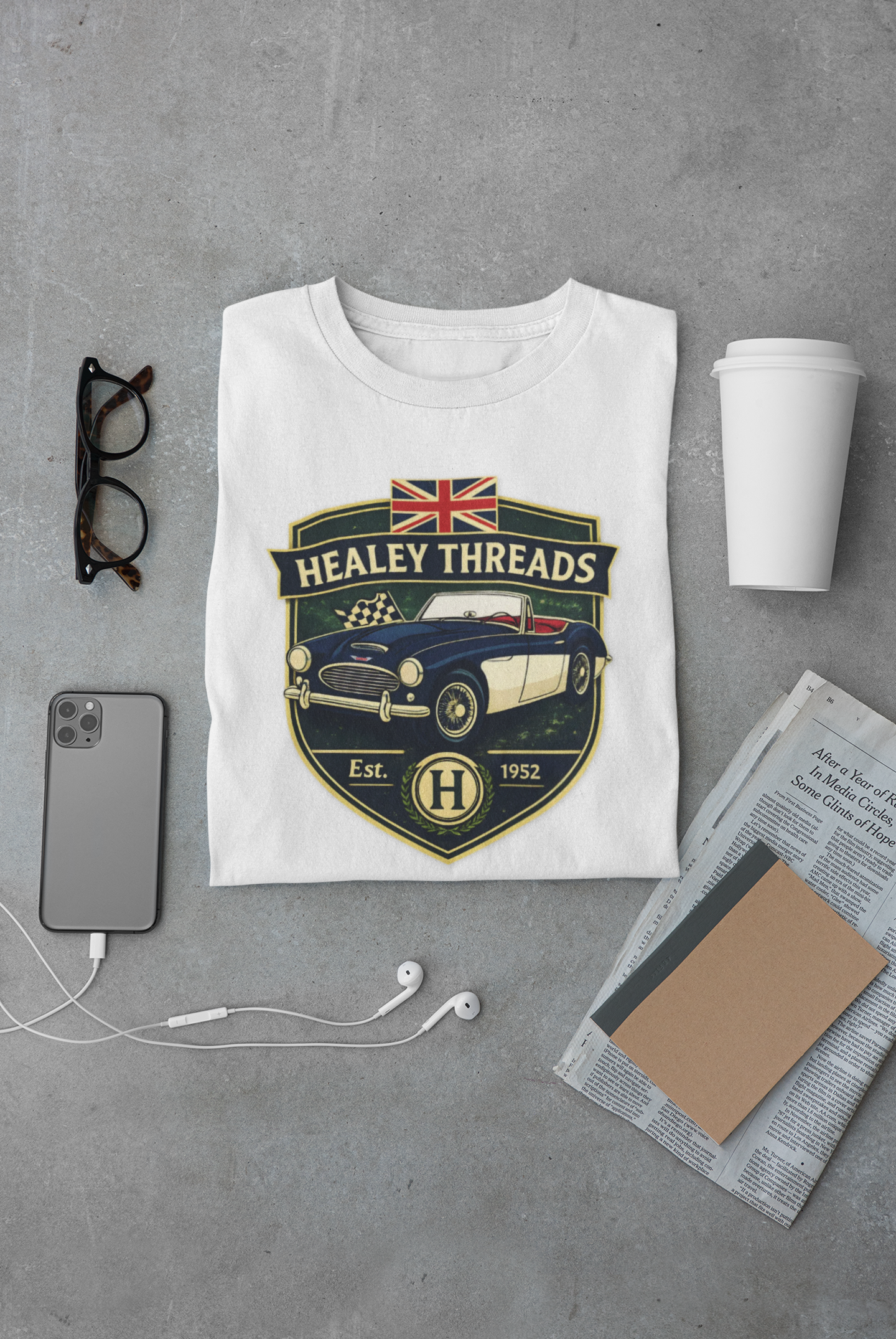 White t-shirt with 'Healey Threads' design on a gray surface with phone, cup, and glasses.