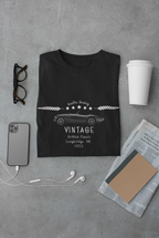 Black t-shirt with vintage car design on a gray surface with phone, earbuds, and coffee cup.