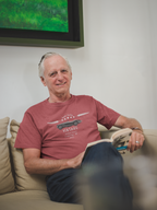 Man sitting on a couch wearing a red t-shirt with a graphic design, in a room with a painting on the wall.